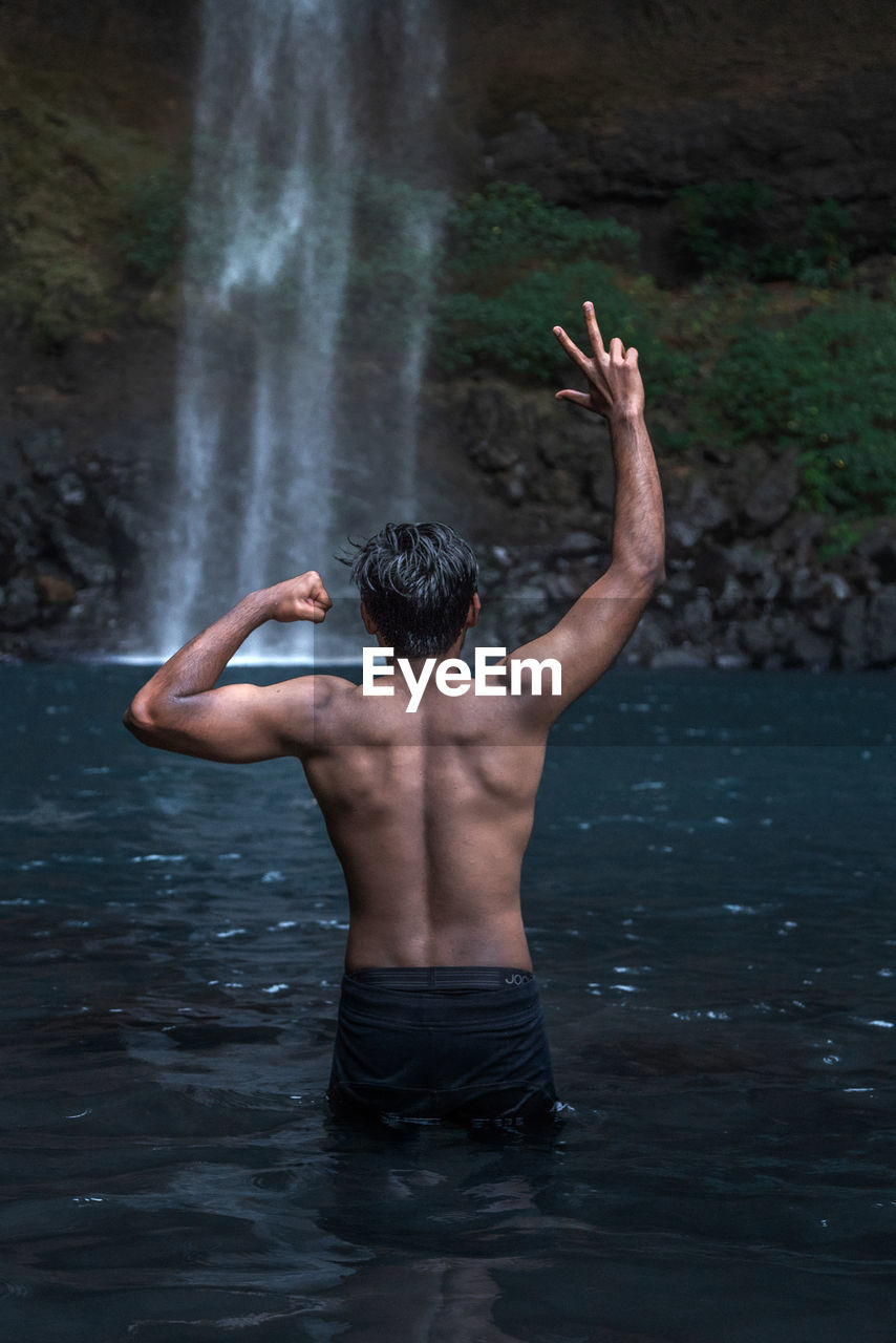 Rear view of shirtless man gesturing while standing in lake by waterfall