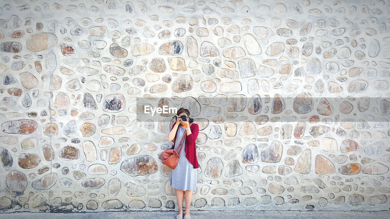 Woman photographing on footpath against wall