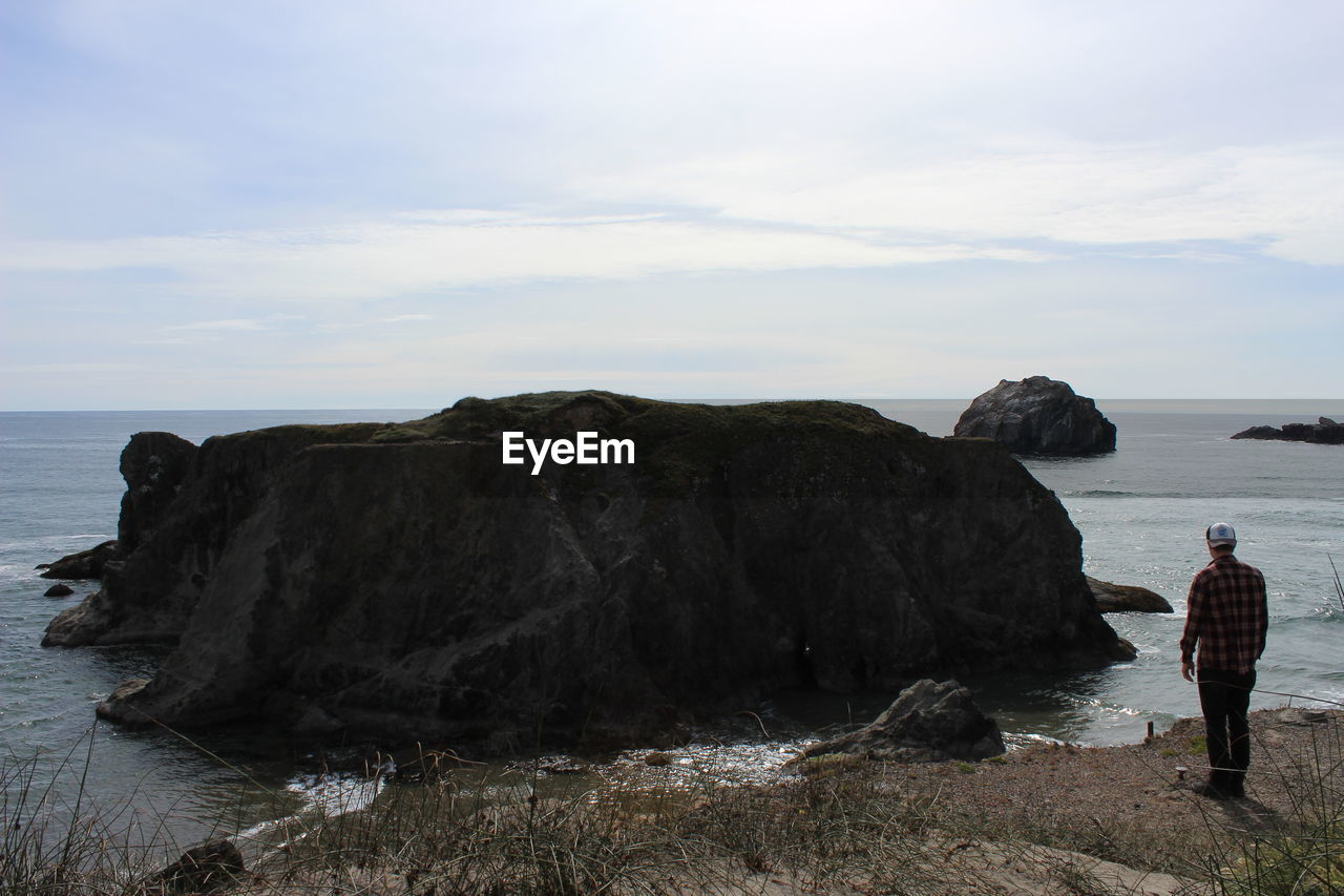 REAR VIEW OF MAN STANDING ON CLIFF BY SEA