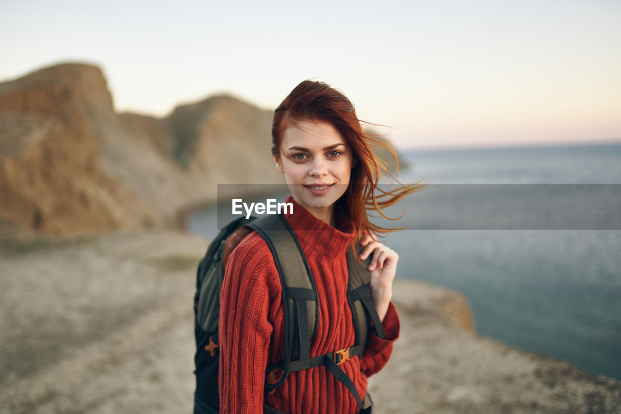 PORTRAIT OF YOUNG WOMAN STANDING ON BEACH DURING SUNSET