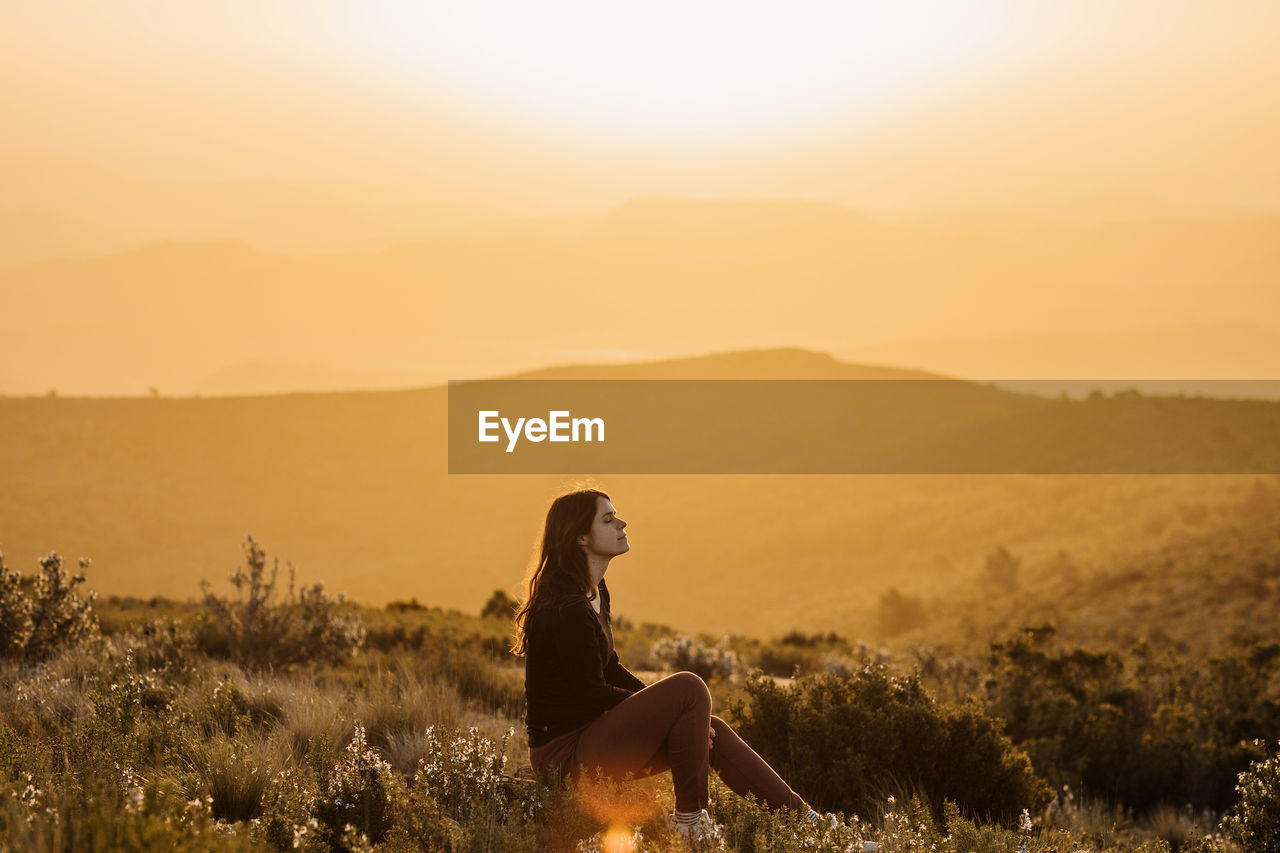 Side view of peaceful female traveler sitting on hill with closed eyes and enjoying nature in highlands at sundown