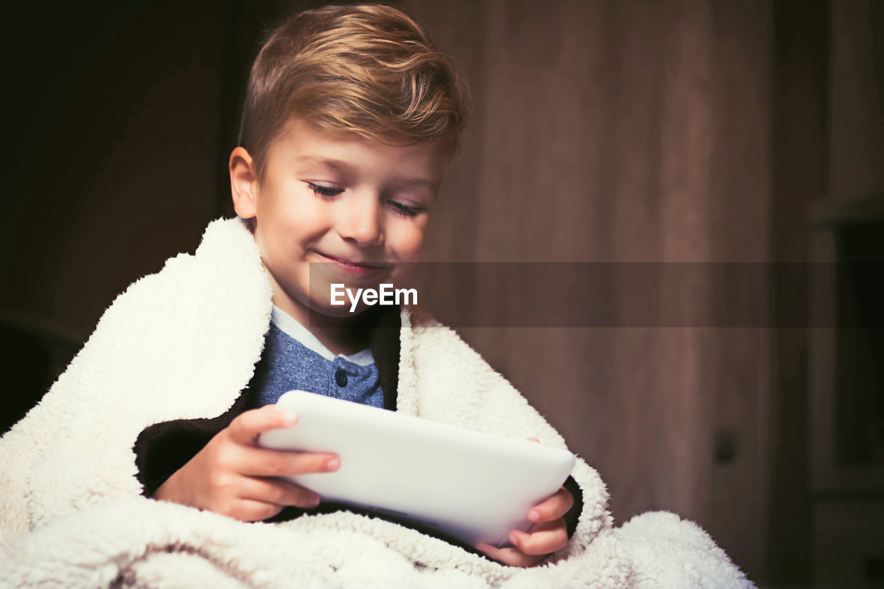 Cute kid covered with blanket using digital tablet in the bedroom.