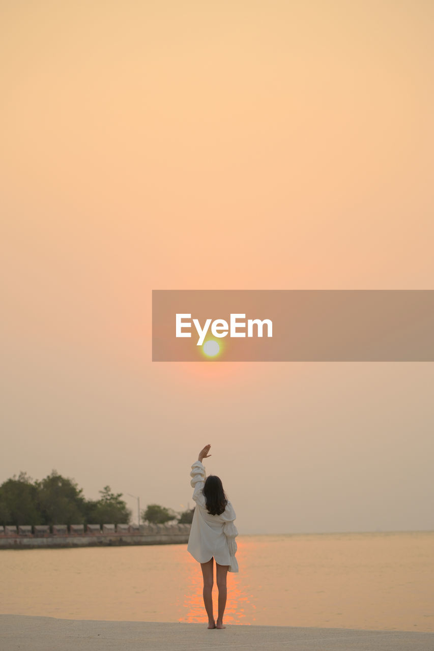 FULL LENGTH REAR VIEW OF WOMAN STANDING ON BEACH DURING SUNSET