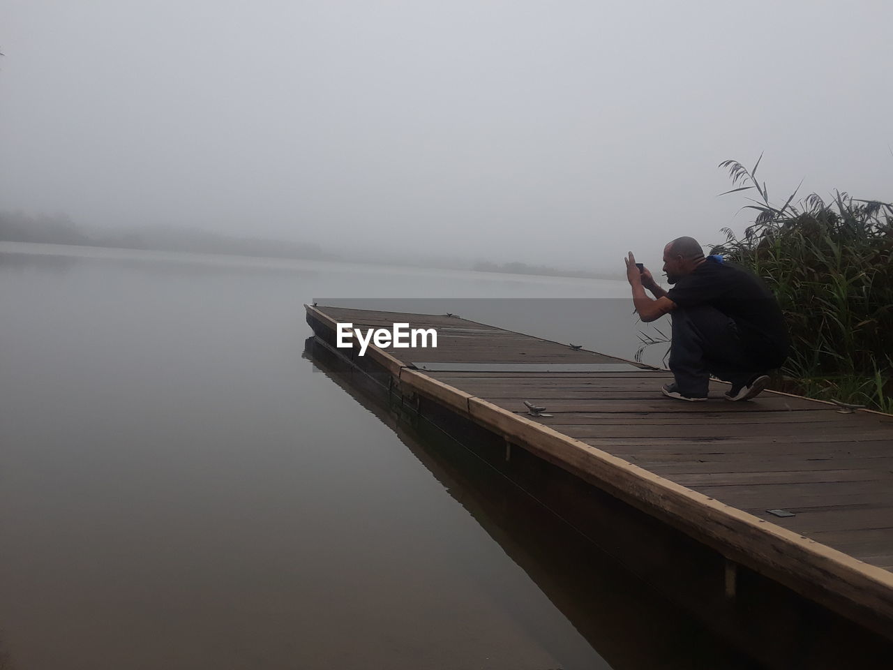 MAN SITTING ON PIER OVER LAKE AGAINST SKY