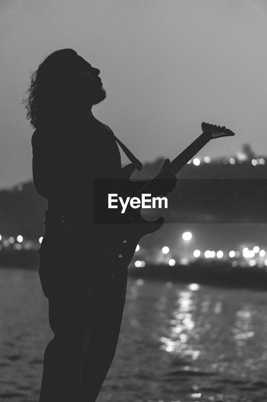 Silhouette of man playing guitar while standing by sea against sky