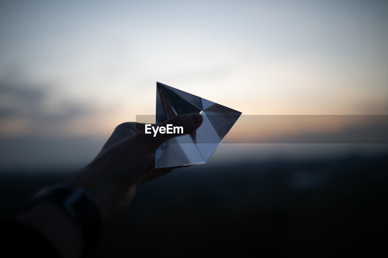 Cropped hand holding crystal against sky during sunset