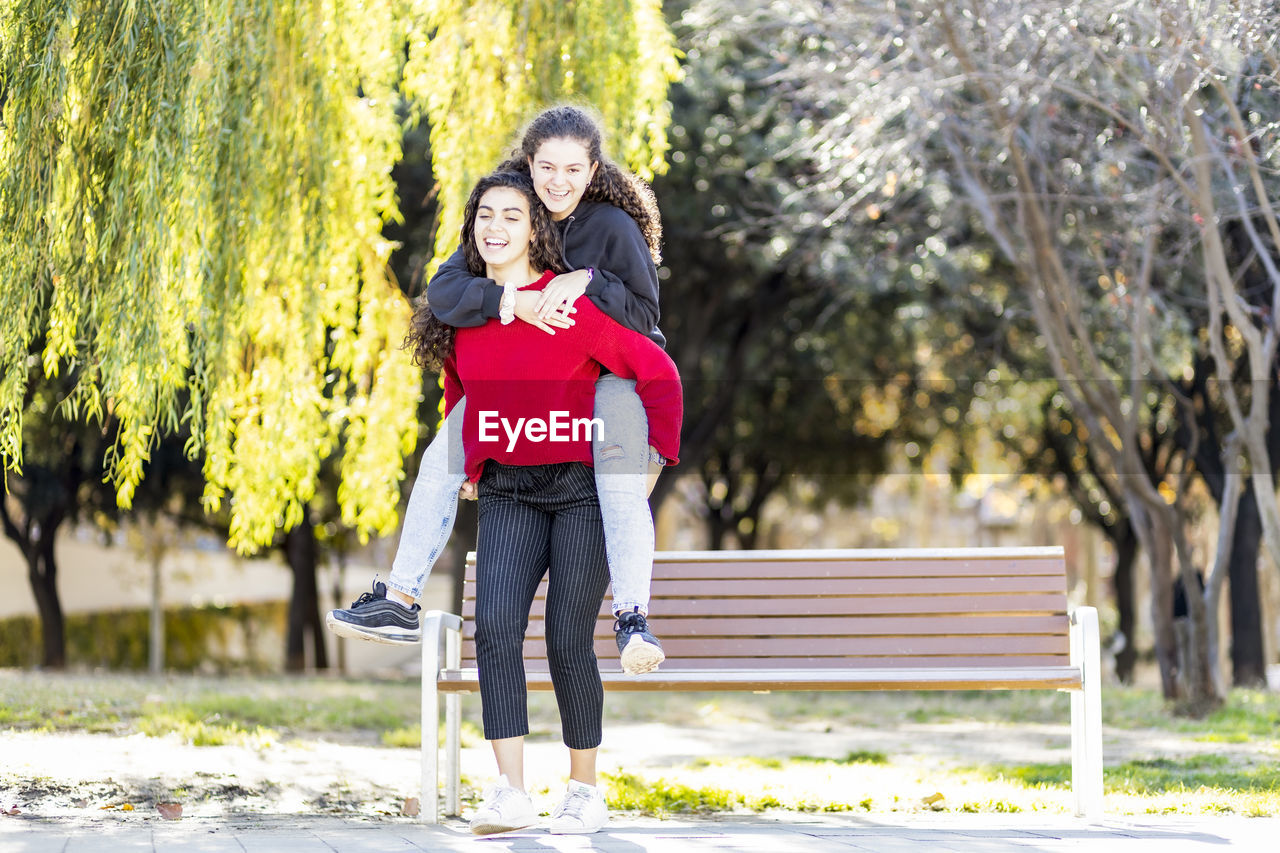 Happy teenage girl giving piggyback ride to friend in park