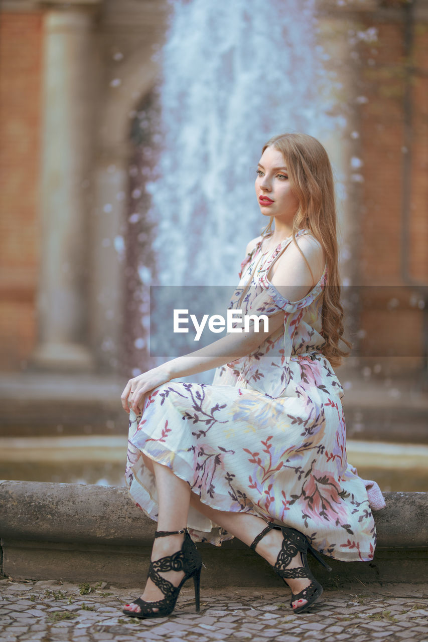 Side view of beautiful young woman sitting against fountain in city