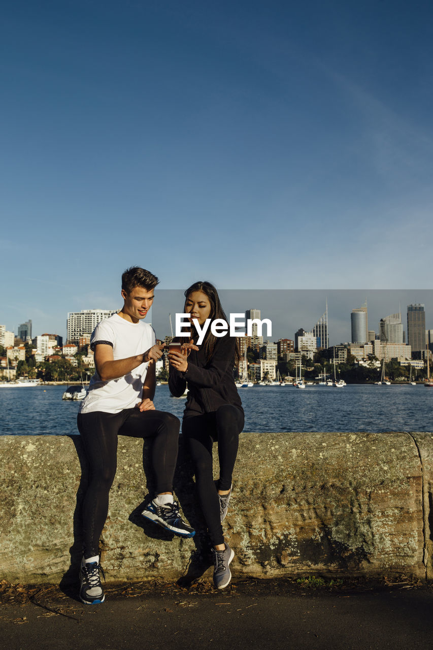 Full length of young couple using smart phone while exercising on promenade against cityscape