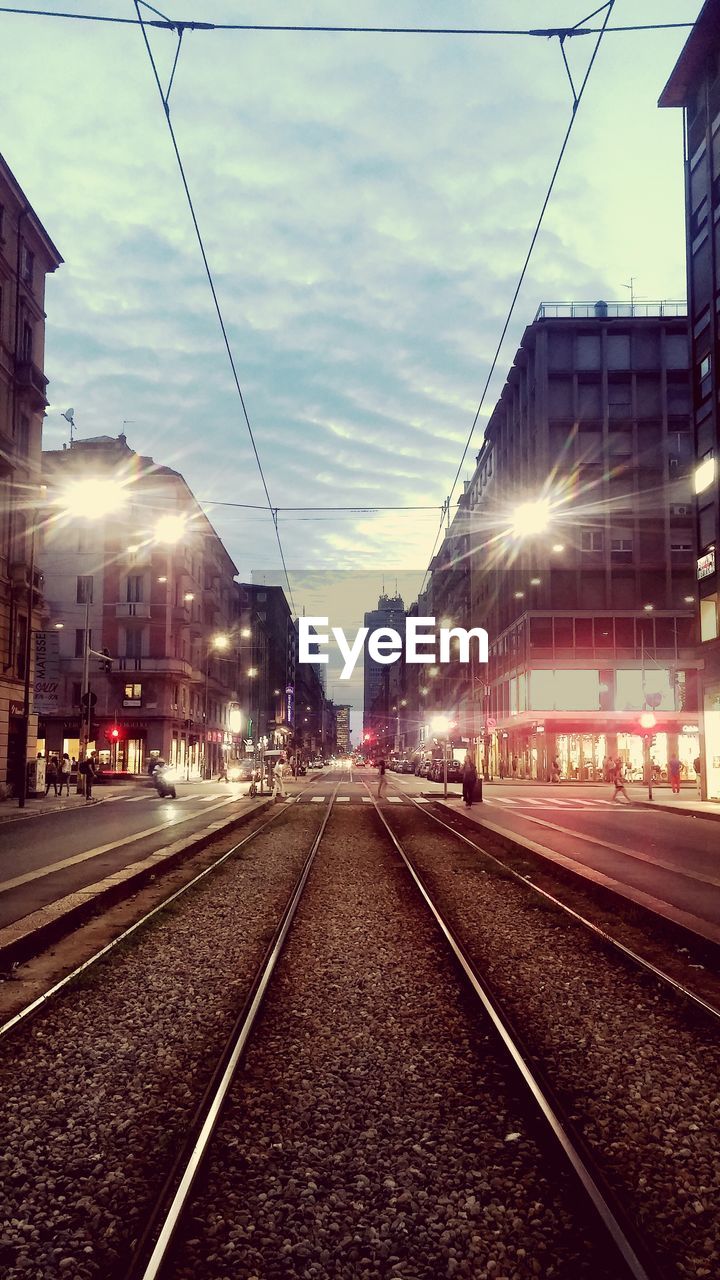 Railroad tracks amidst illuminated buildings against cloudy sky at dusk