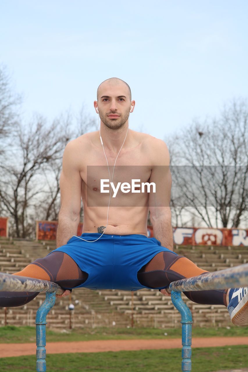 Portrait of shirtless man listening music while exercising against sky