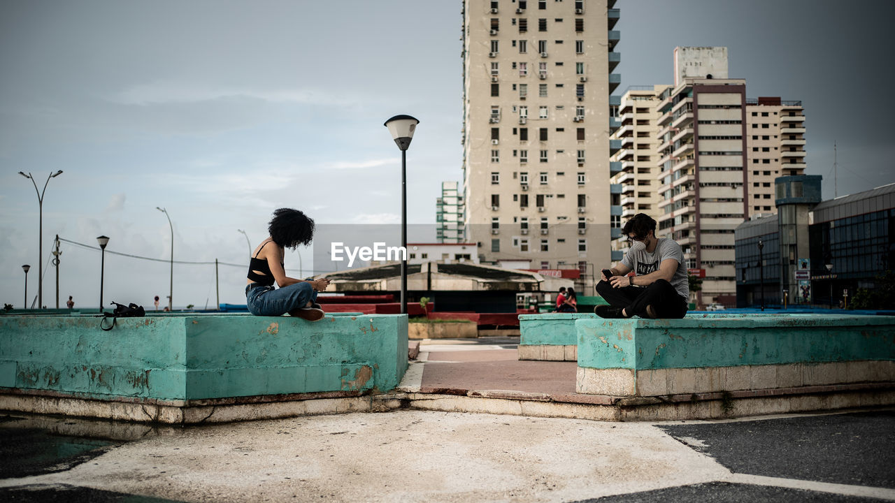 PEOPLE SITTING ON STREET AMIDST BUILDINGS IN CITY