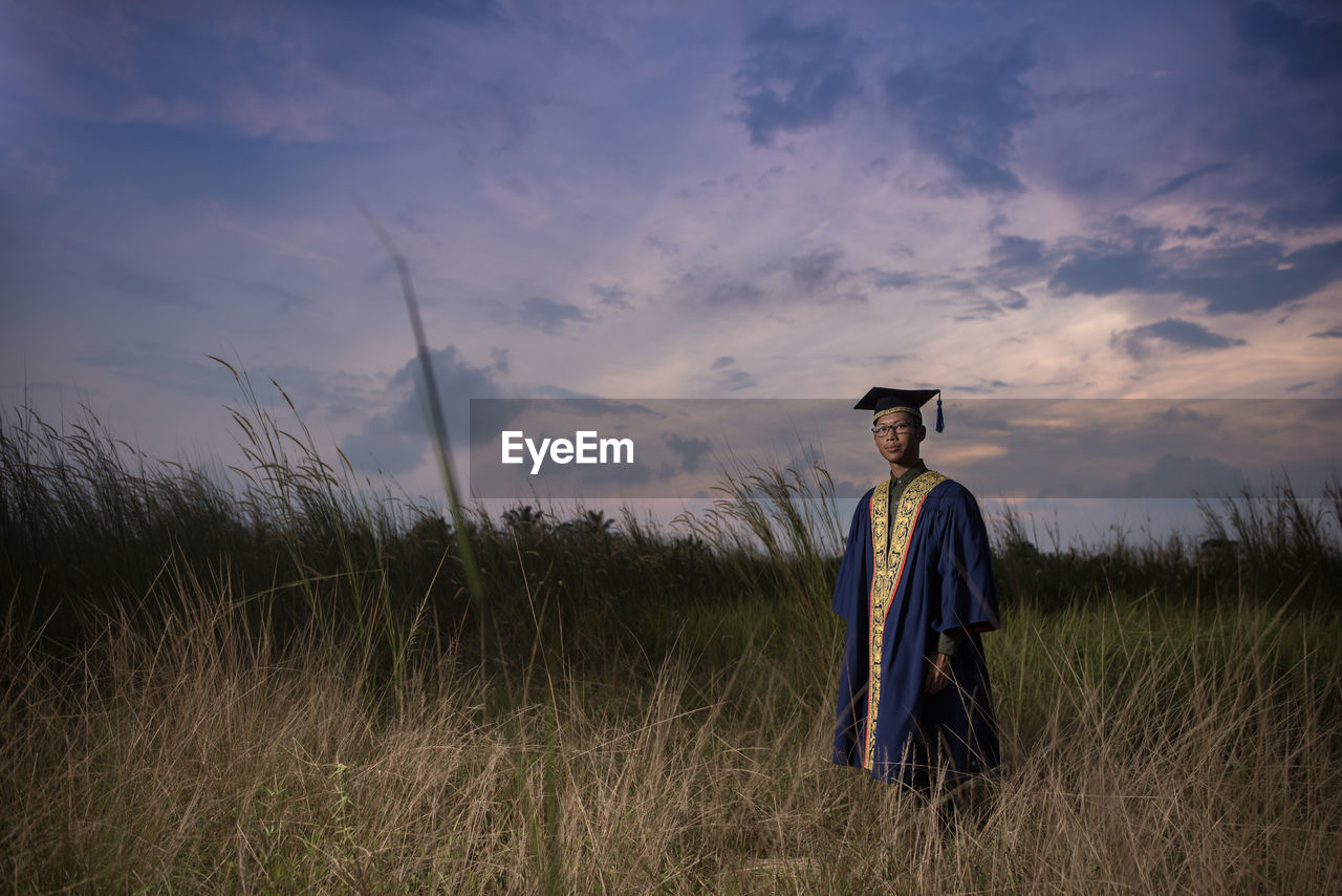 Man in graduation gown standing on field against sky