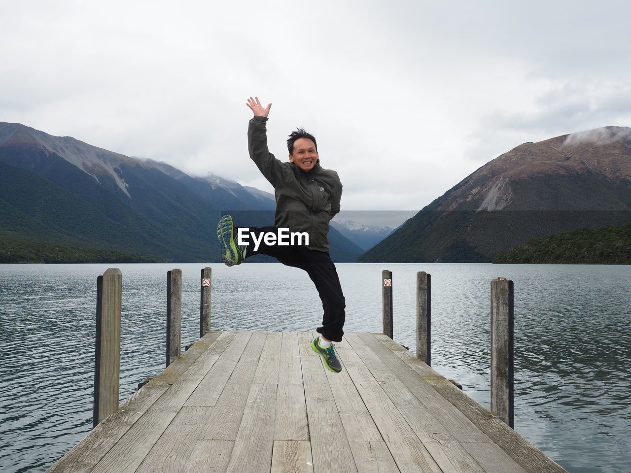 Portrait of cheerful man jumping on pier over lake against mountains