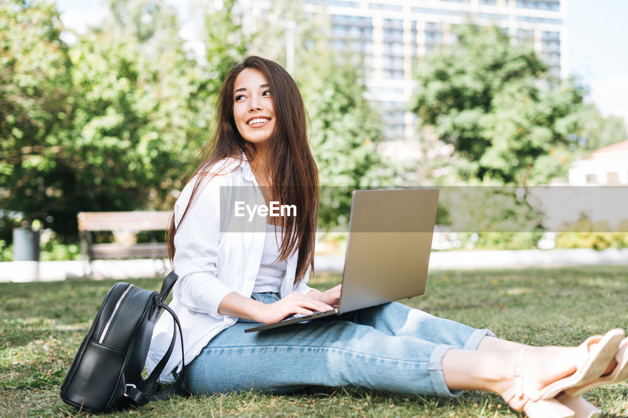 Young asian woman student freelancer with long hair working on laptop in city park