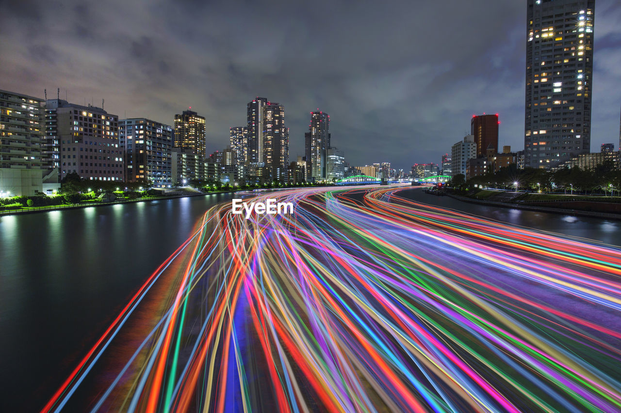 Light trails on city by buildings against sky at night