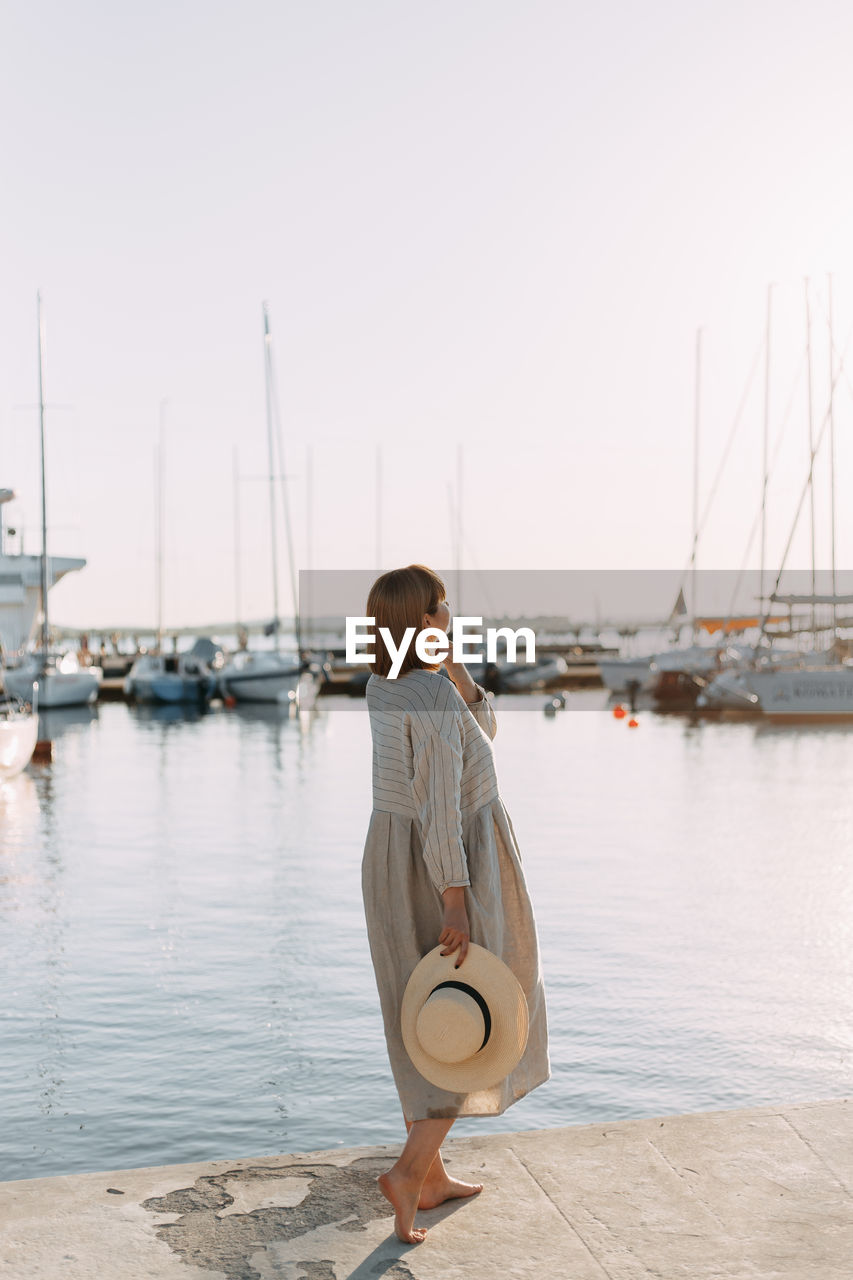 Happy young woman in dress and hat walks on the beach near the boats