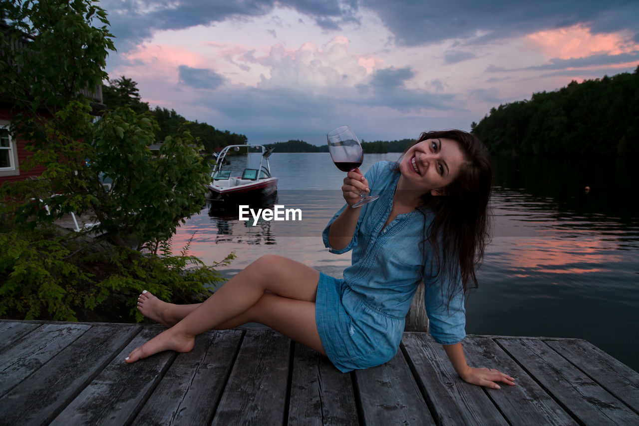 YOUNG WOMAN SITTING ON WOOD AGAINST SKY DURING SUNSET