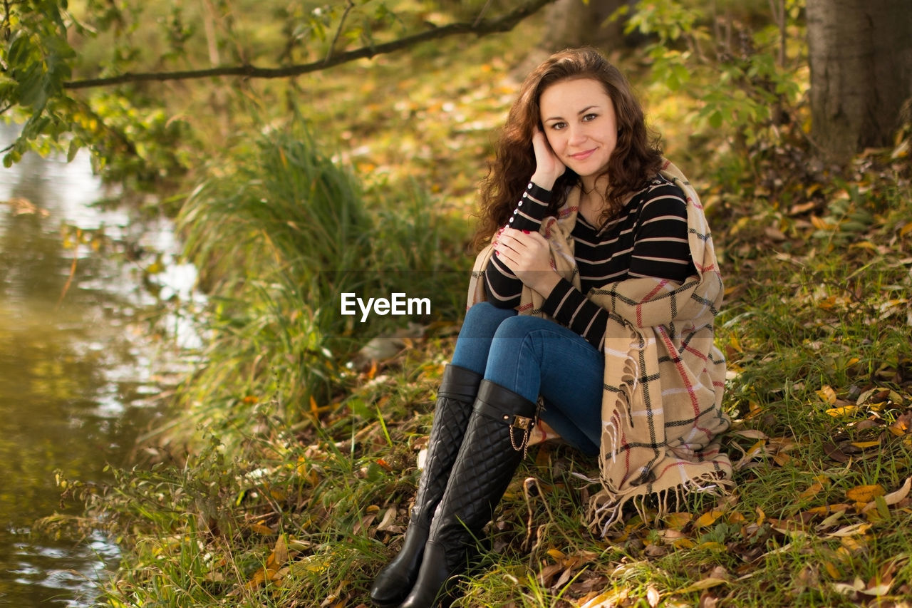 Full length of young woman smiling while sitting at grassy lakeshore