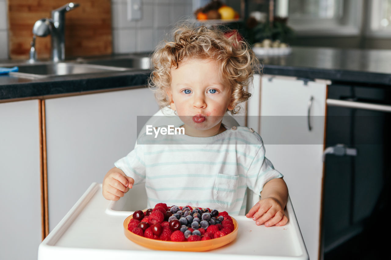  caucasian baby boy eating red fruits at home. funny child kid sitting high chair with fresh berries 