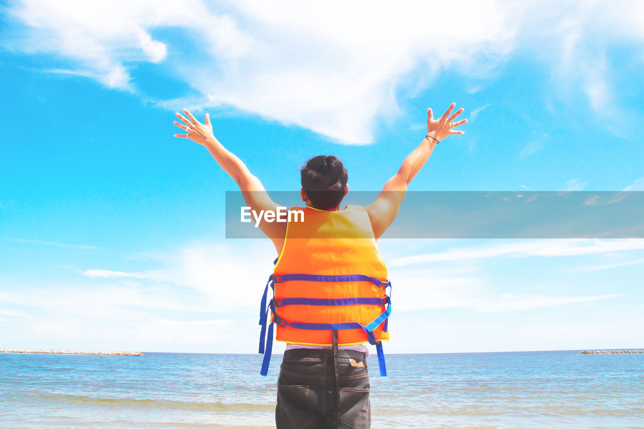 Rear view of man with arms raised standing at beach against sky