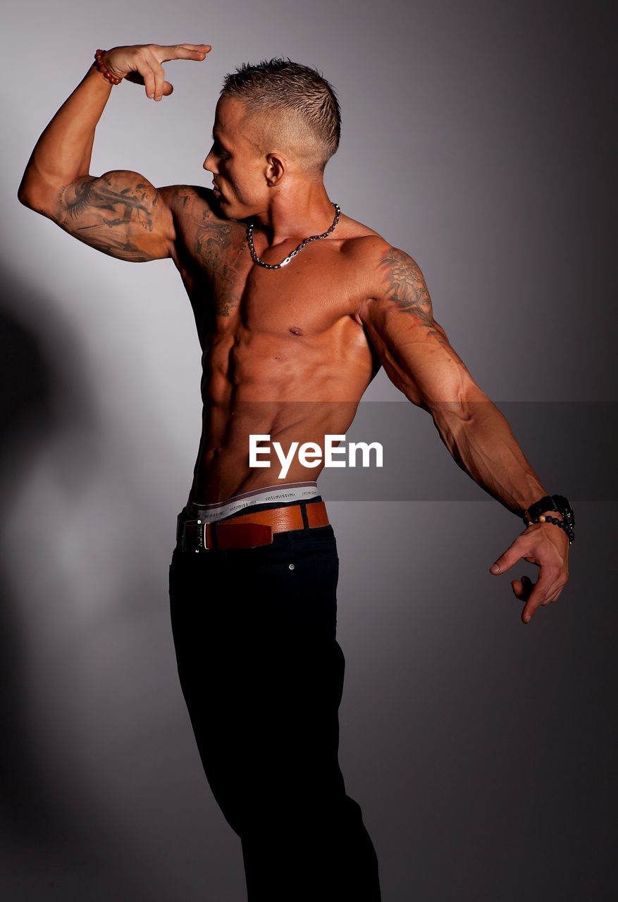 Shirtless young man flexing muscles while standing against wall