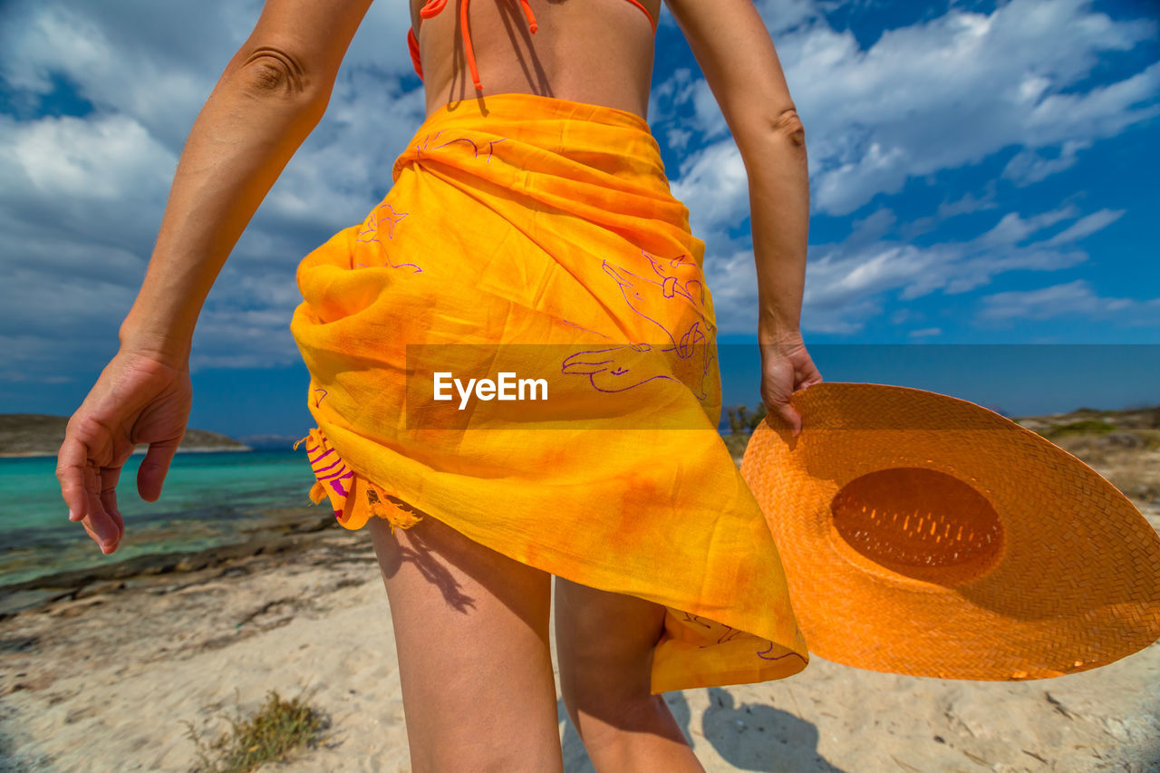 Midsection of woman in yellow sarong holding hat at beach against sky