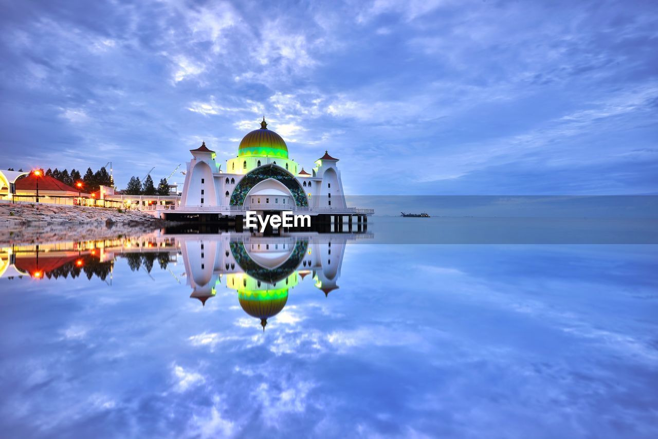 Illuminated mosque reflecting on calm sea against cloudy sky during sunset