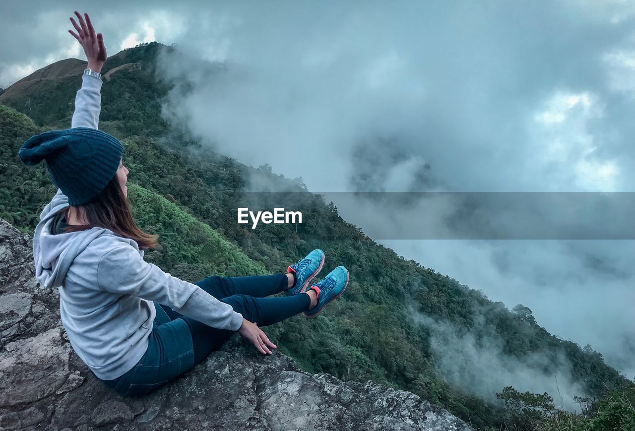 Full length of carefree young woman sitting on cliff during foggy weather