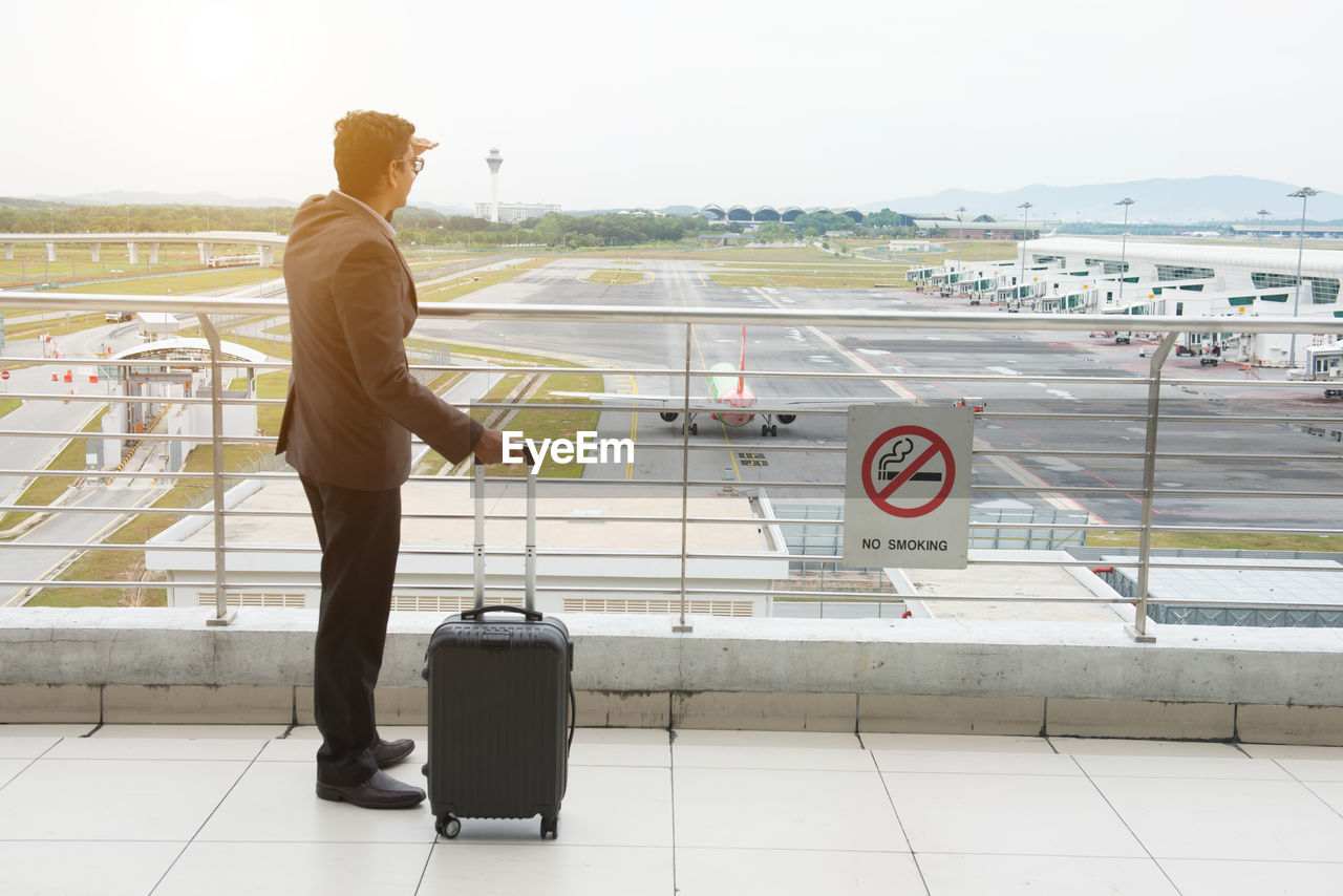 REAR VIEW OF MAN STANDING ON AIRPORT AGAINST SKY