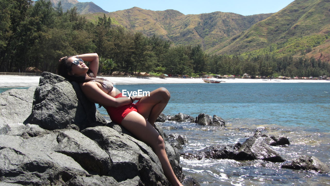 Sensuous woman in bikini leaning on rock formation by lake against mountains