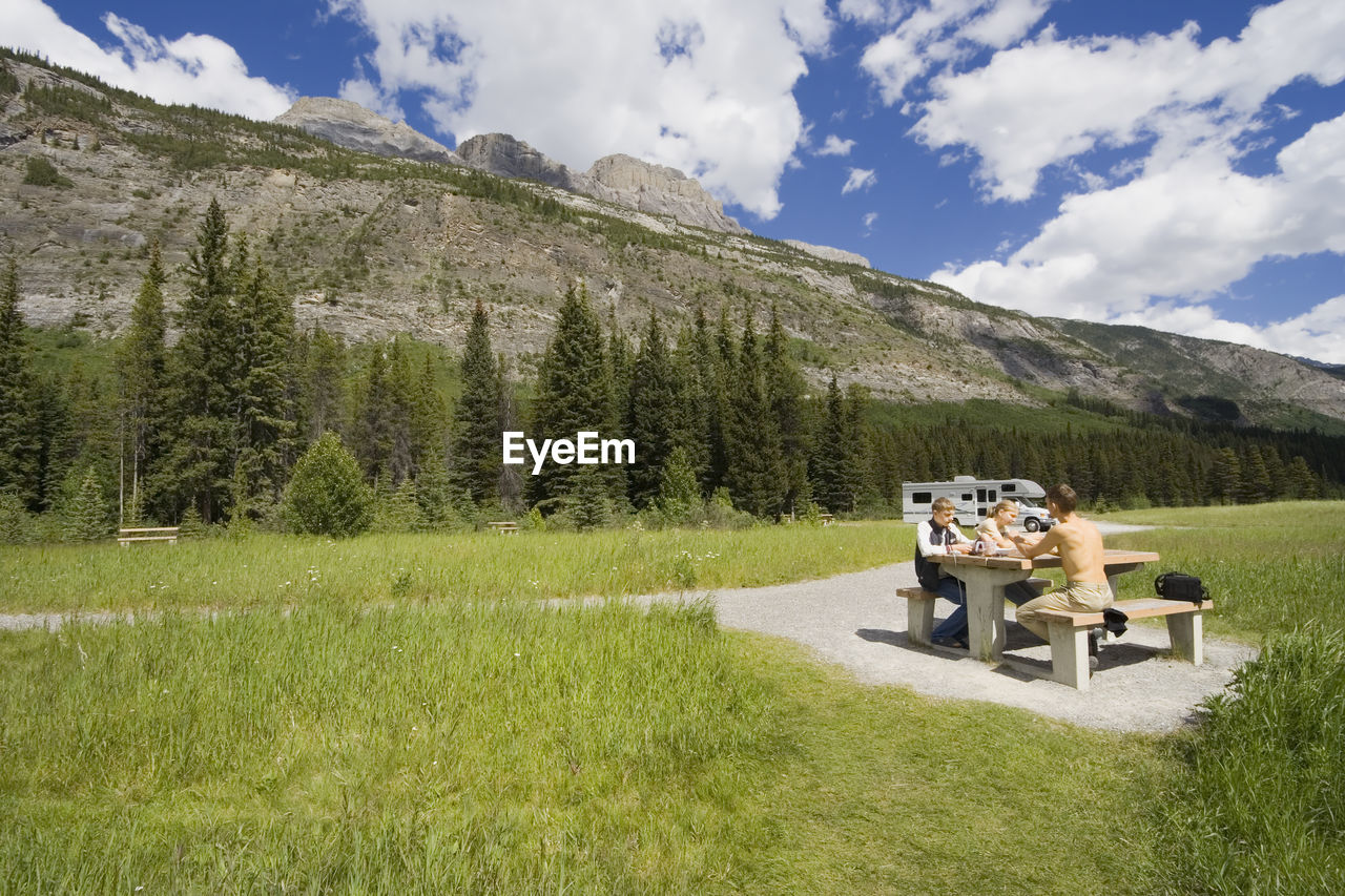 Family sitting on picnic table by mountain against cloudy sky