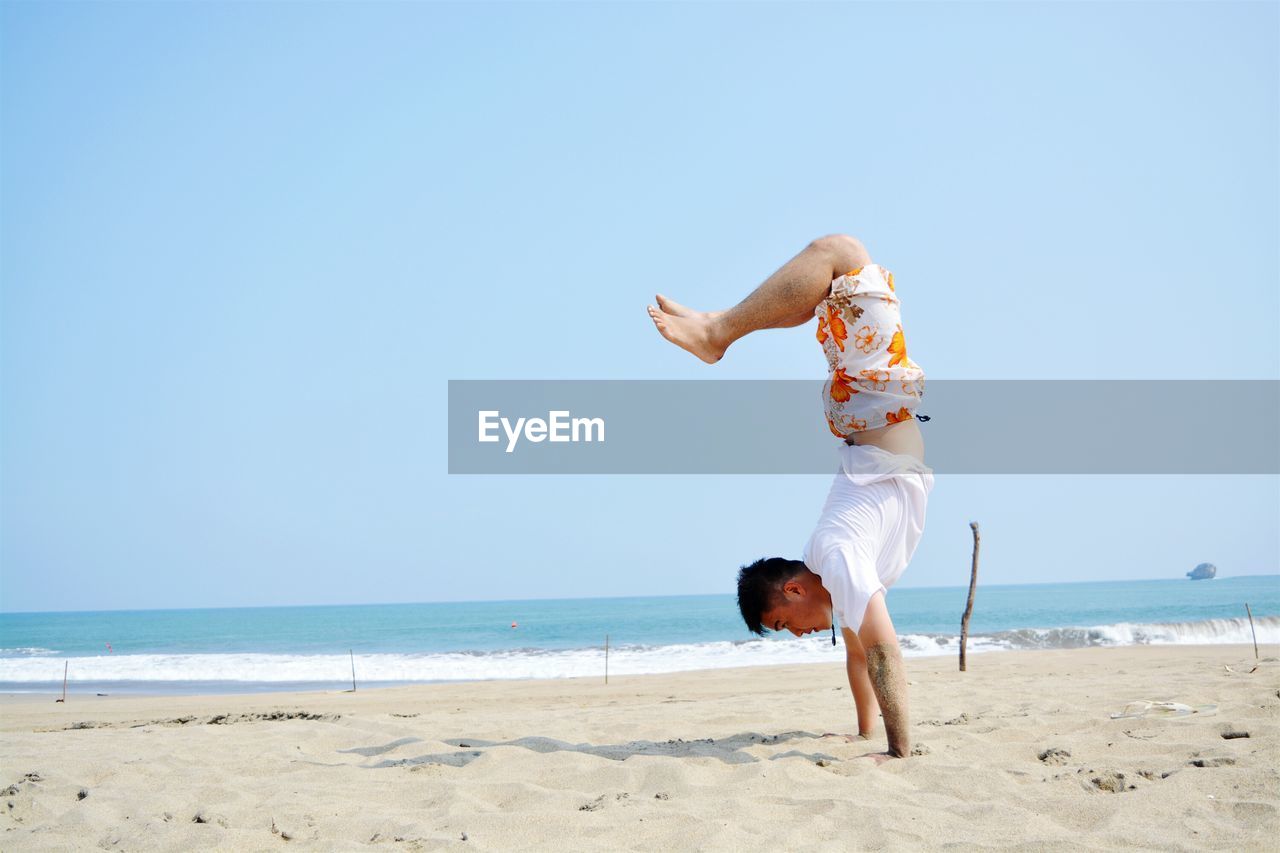 Side view of man doing handstand at beach against clear blue sky during sunny day