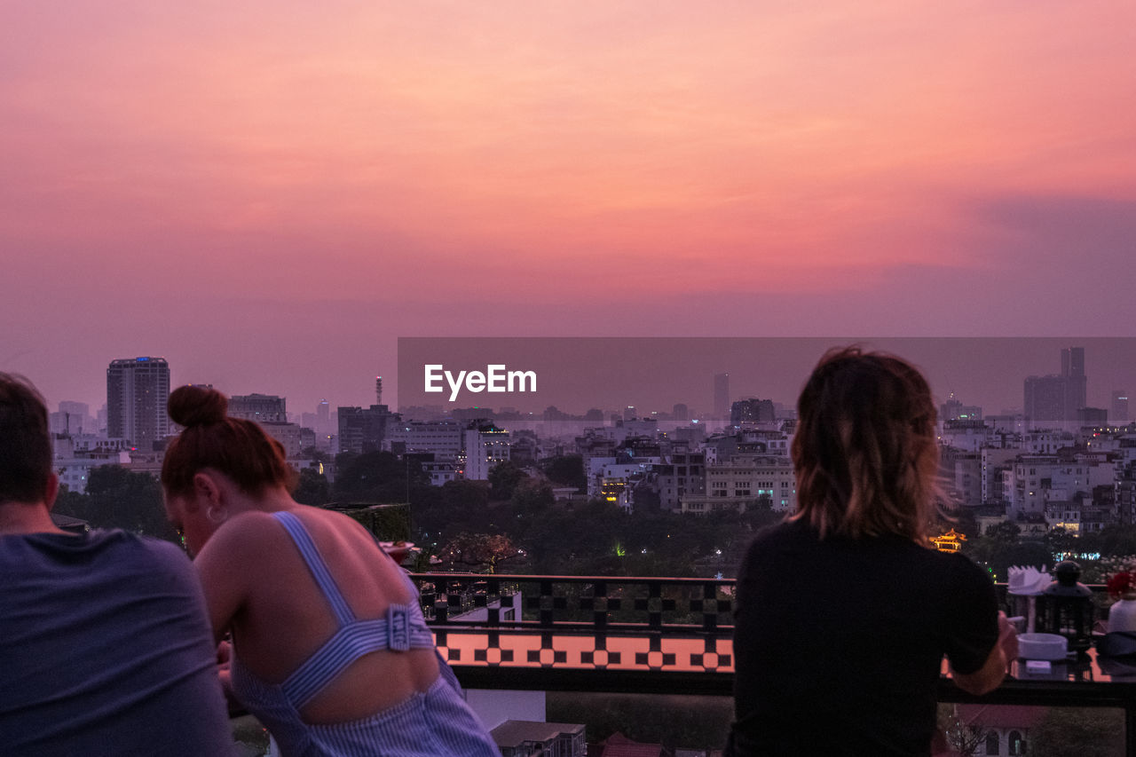 REAR VIEW OF PEOPLE LOOKING AT CITYSCAPE AGAINST SKY