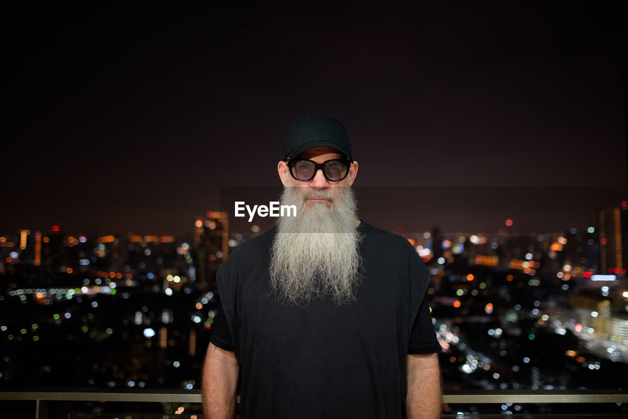 Portrait of man standing against illuminated cityscape at night