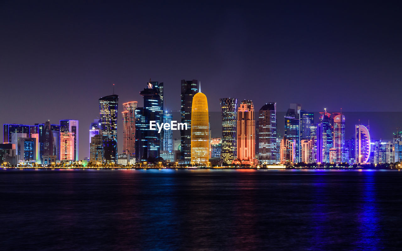 Illuminated doha tower and skyscrapers by river in city against clear sky at night