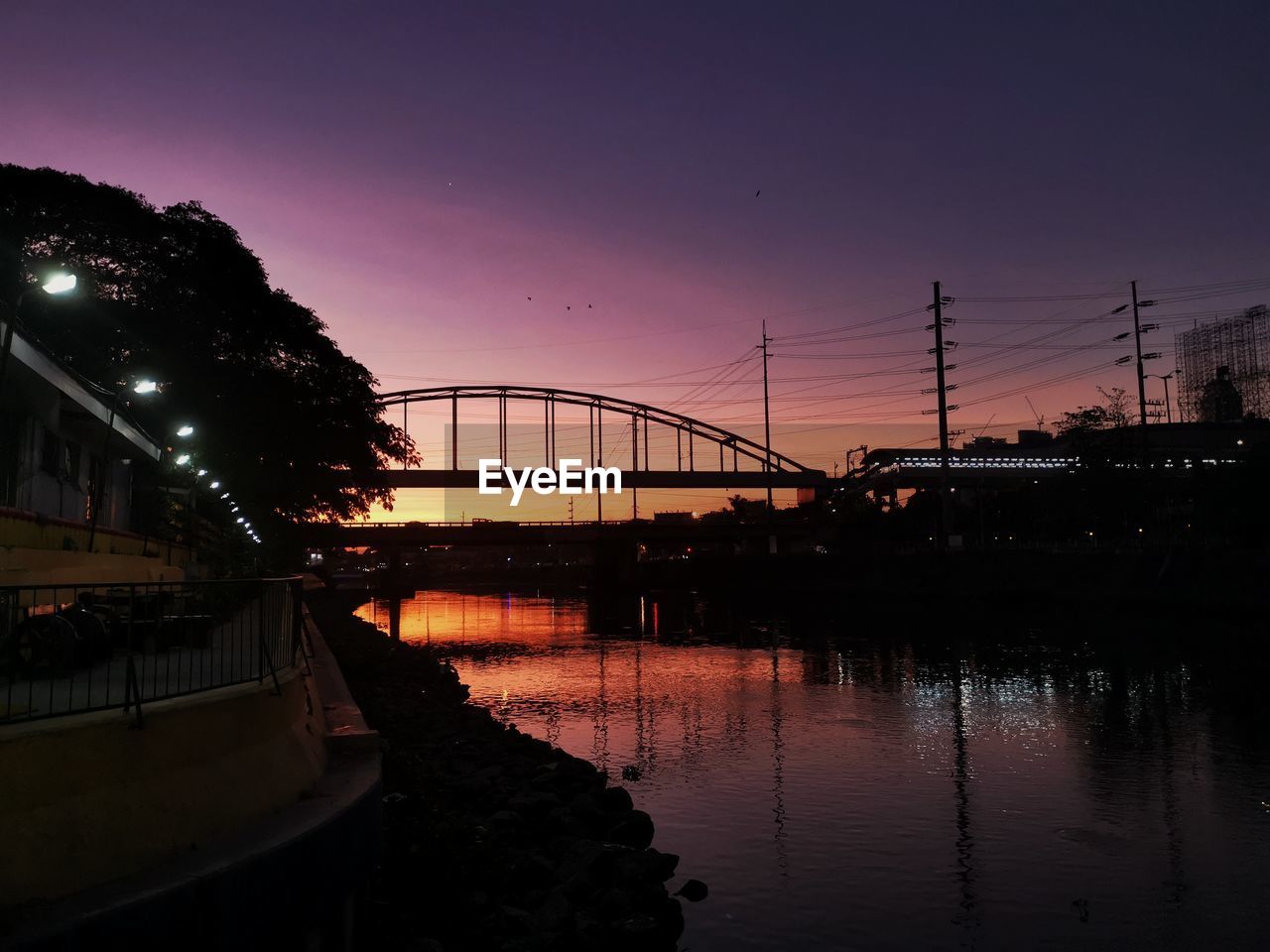 BRIDGE OVER RIVER IN CITY AGAINST SKY DURING SUNSET