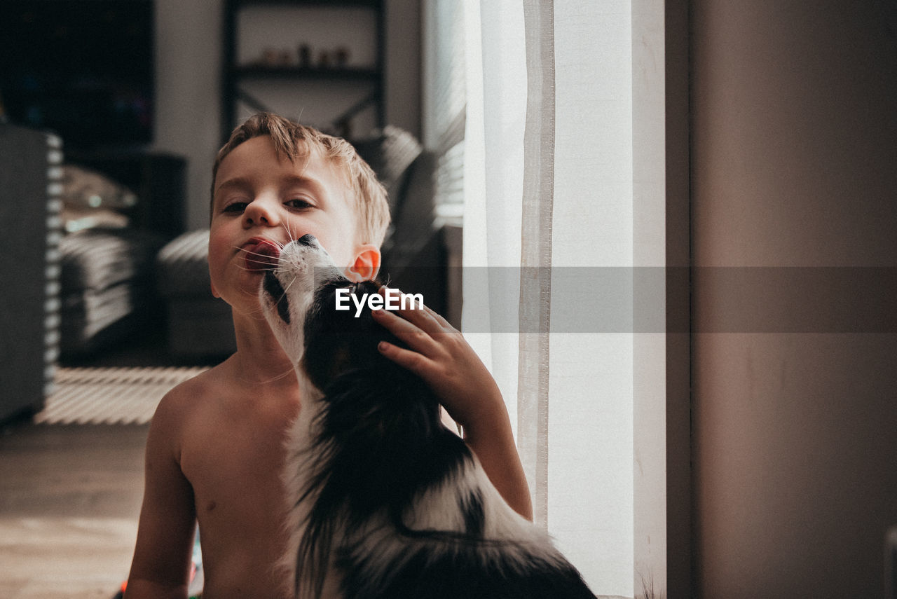 Portrait of shirtless boy kissed by dog at home