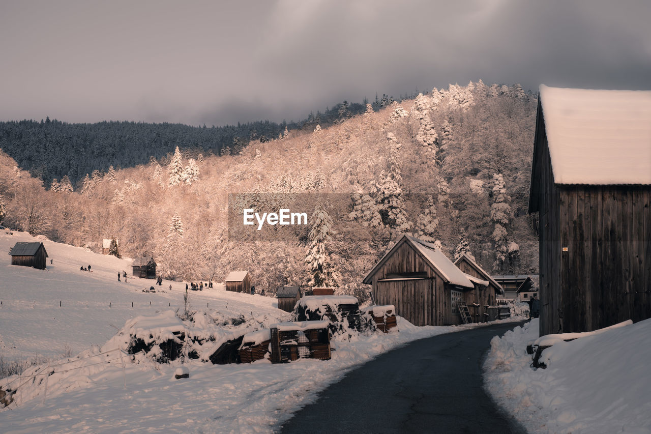 Road leading to snow covered houses by trees against mountain anf sky during winter 