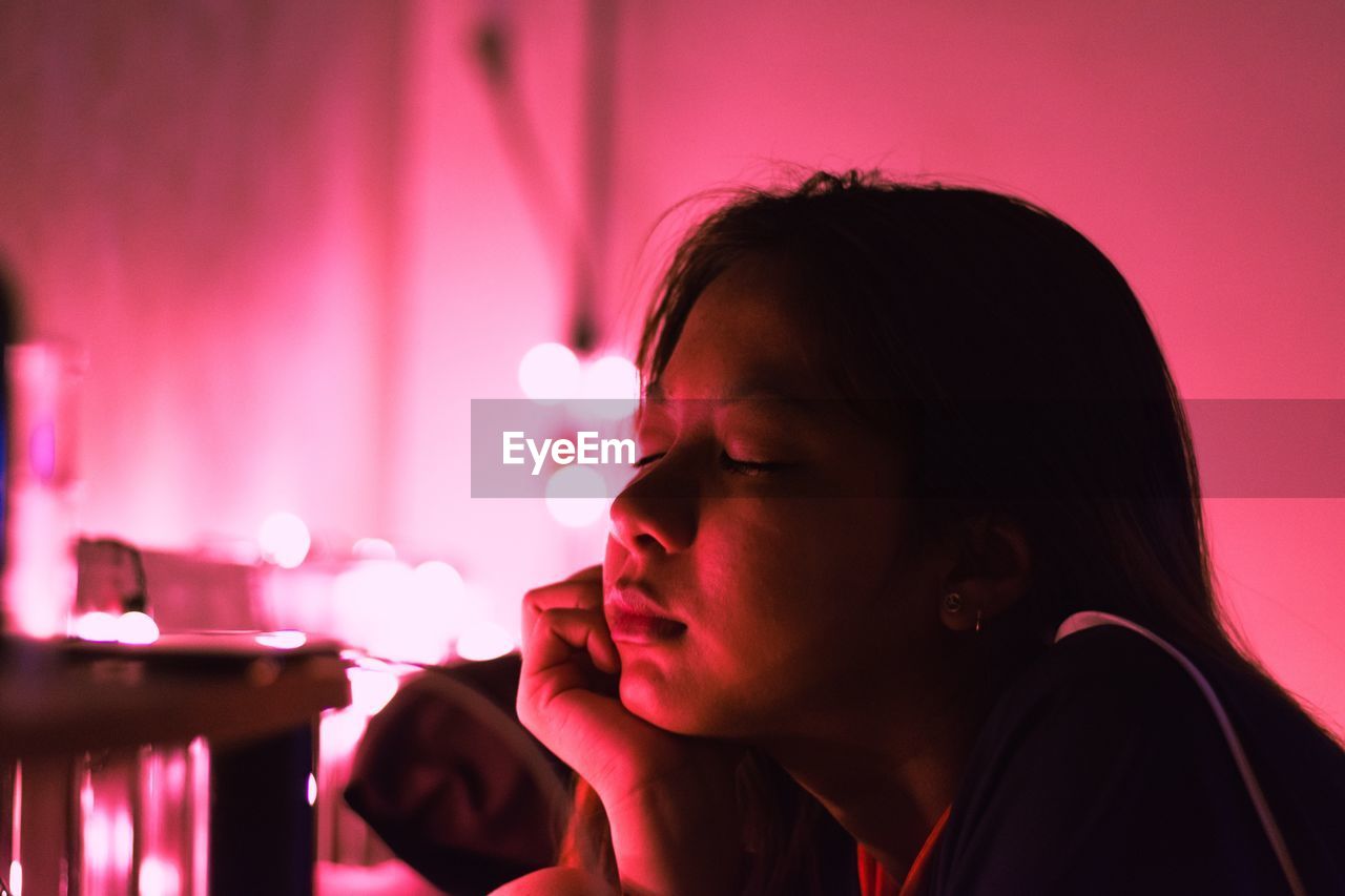 Close-up of young woman with eyes closed lying against pink light at home