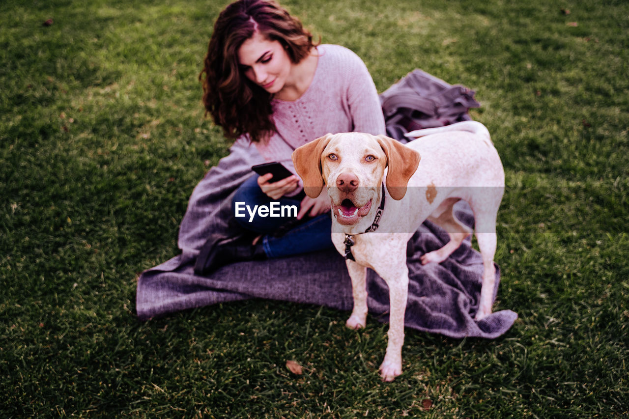 Smiling woman using phone while sitting with dog at park