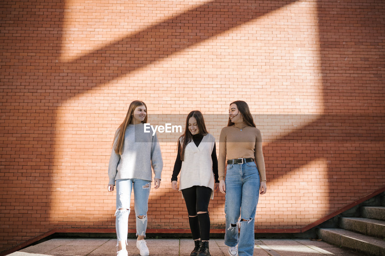 Smiling female friends walking on footpath against brick wall in city