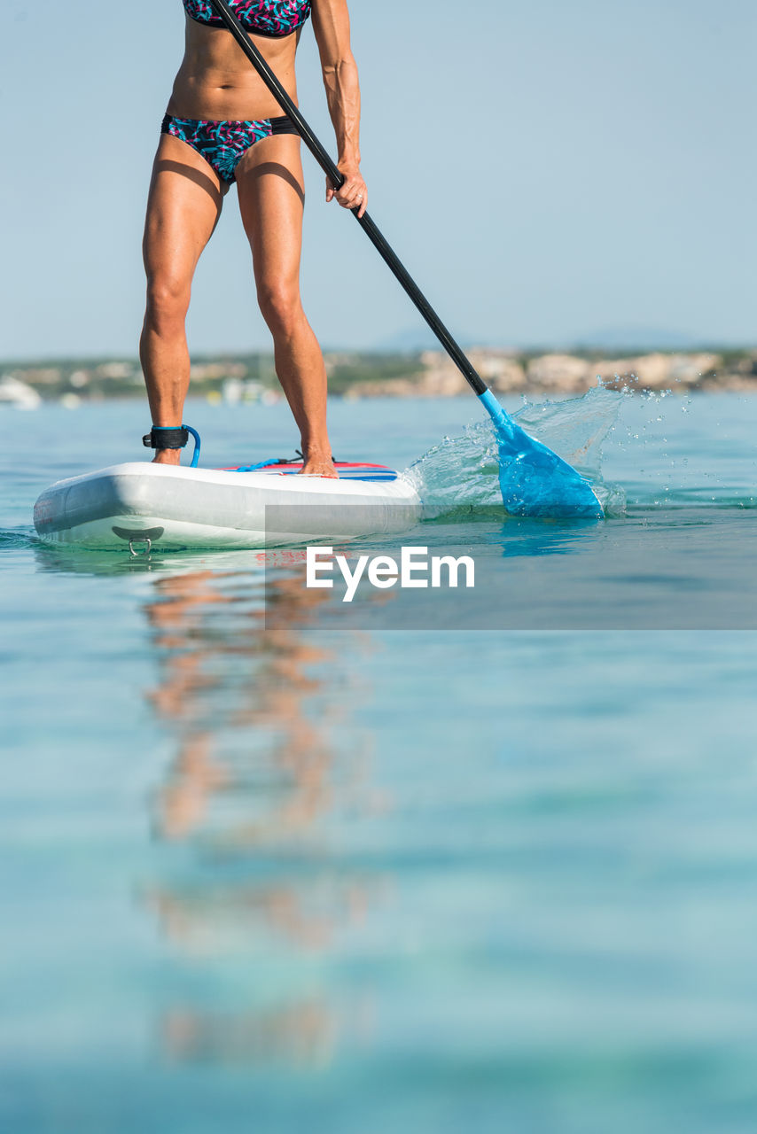 Crop anonymous female surfer standing on surfboard and rowing with paddle while practicing on surface of sea in summer