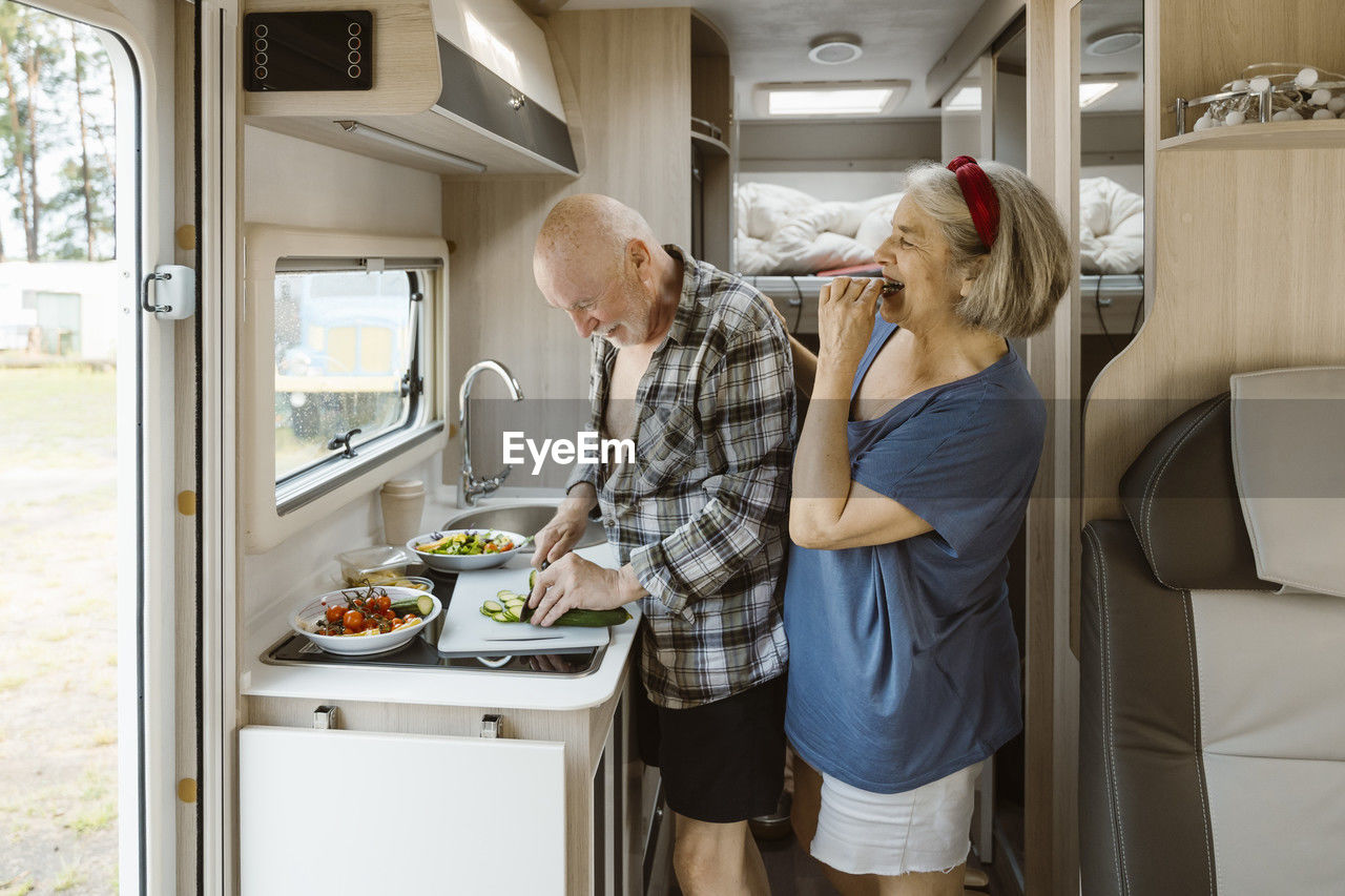 Happy senior woman standing behind man cutting cucumber in motor home