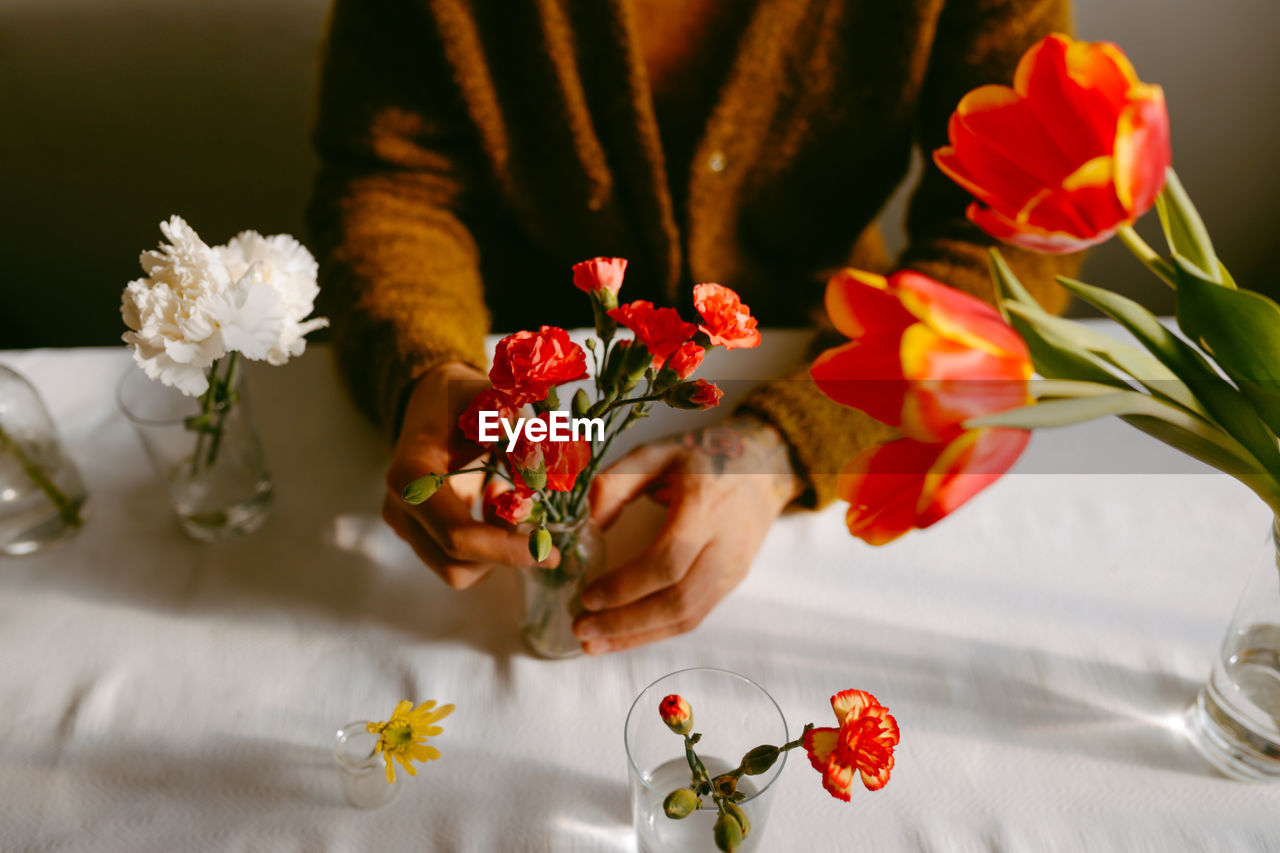 High angle of crop anonymous male florist sitting at table with carnations and tulips in glassware