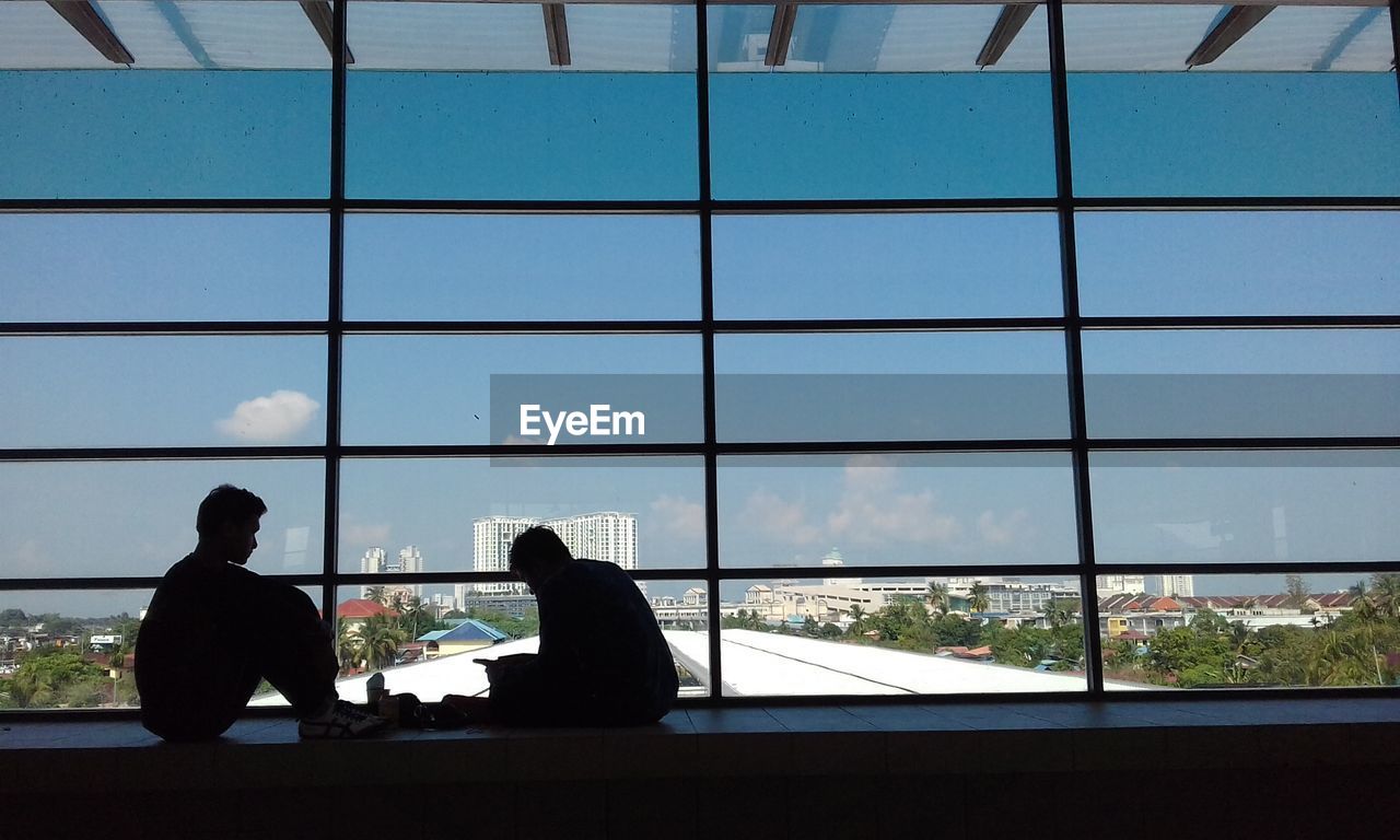 Silhouette men sitting by window in city against sky