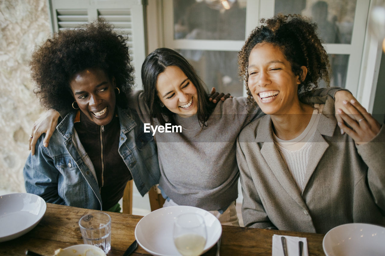 Cheerful woman sitting with arm around female friends at dining table enjoying dinner in patio