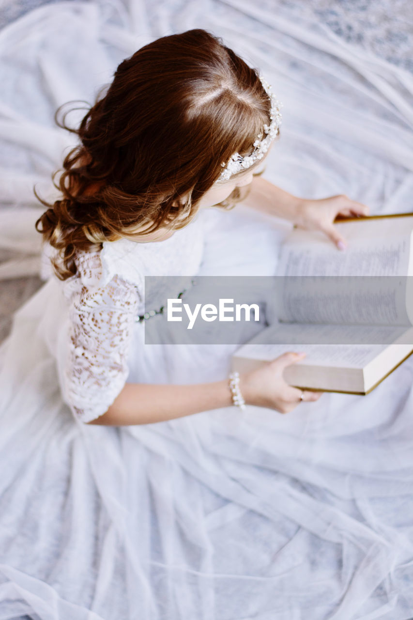 High angle view of girl reading book while sitting on bed at home