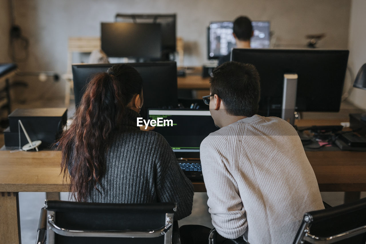 Rear view of young male and female colleagues coding together on laptop at startup company