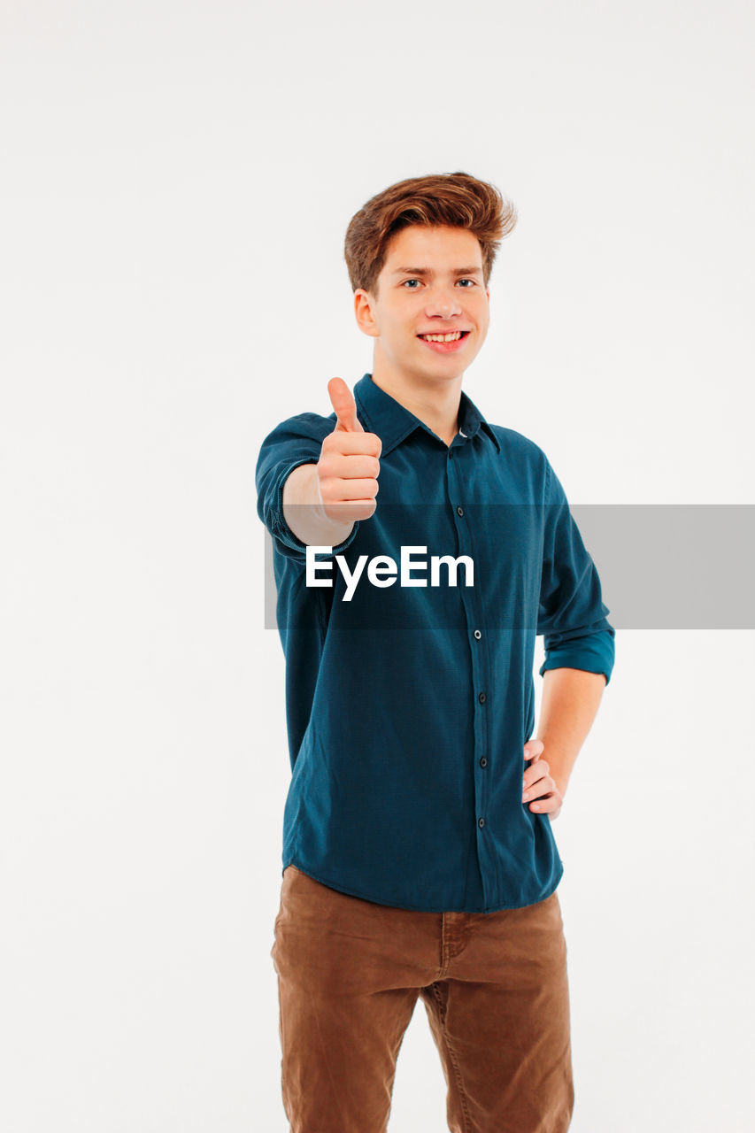 Portrait of young man showing thumbs up while standing against white background